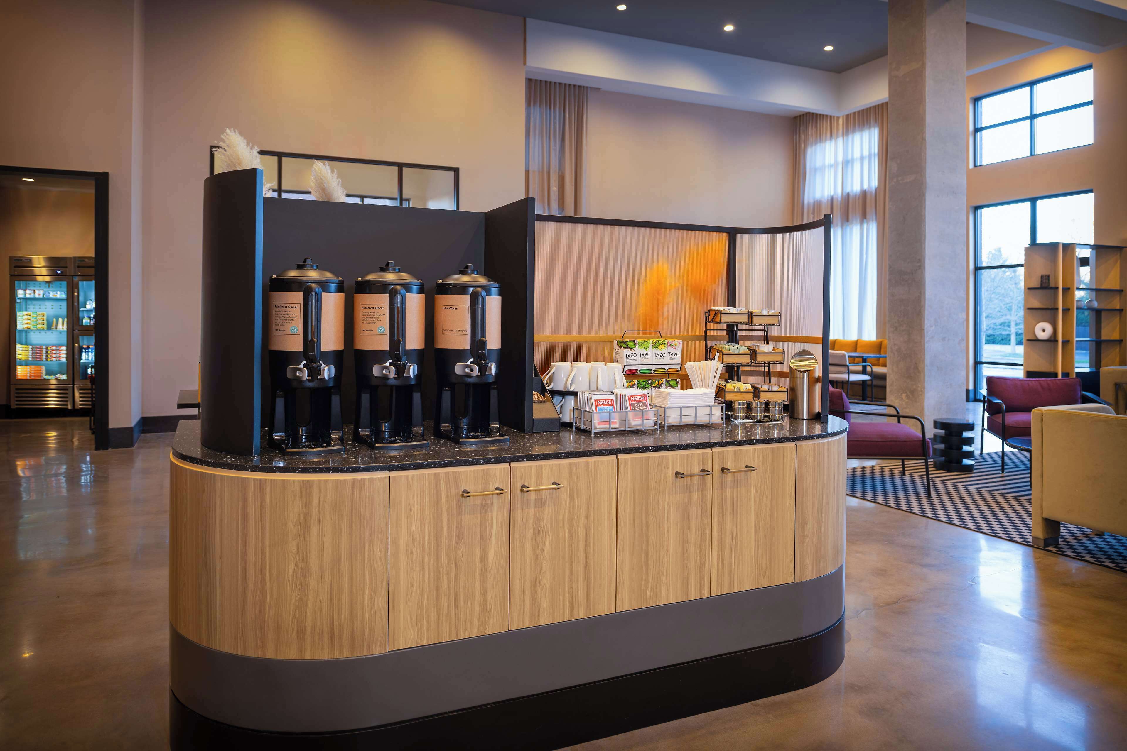 Una moderna zona de recepción de hotel con un mostrador de madera, máquinas de café y un espacio de estar con sillas rojas.