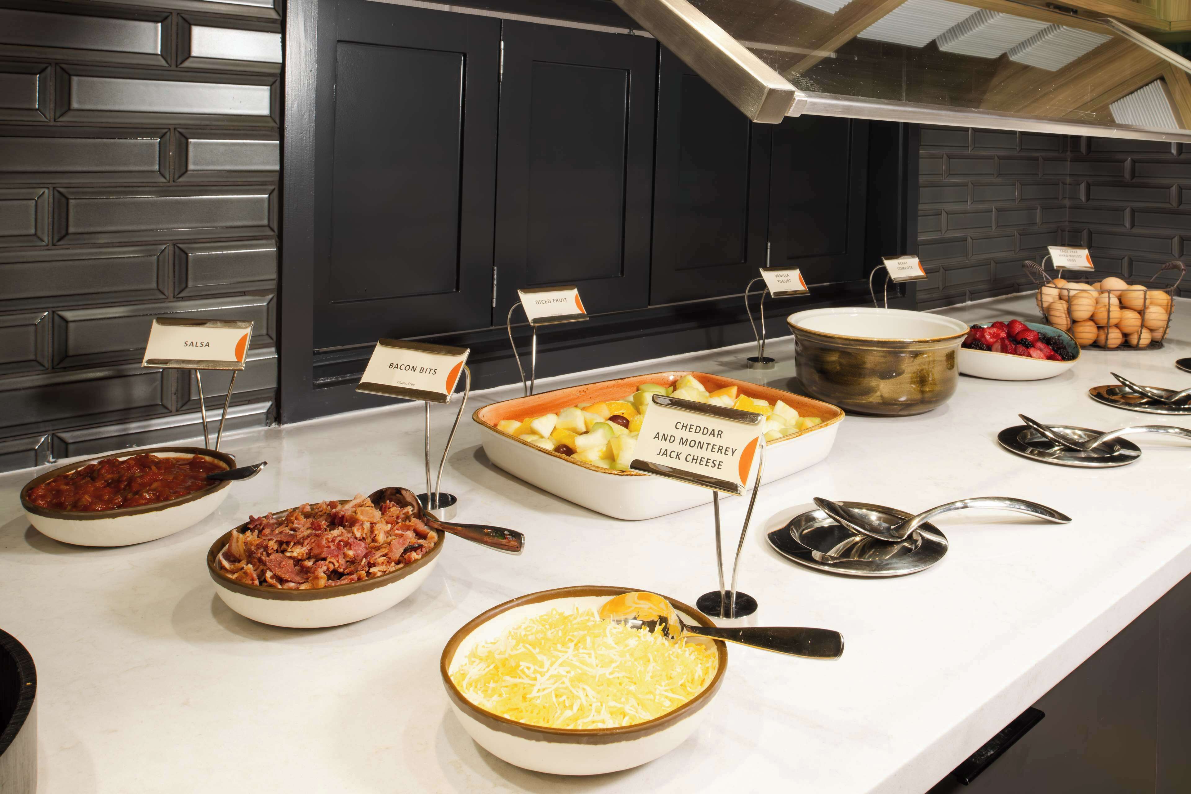 Un buffet proposant divers plats, chacun étant clairement identifié, notamment des « salsa », des « morceaux de bacon », du « fromage cheddar et Monterey Jack » et des « fruits coupés ».