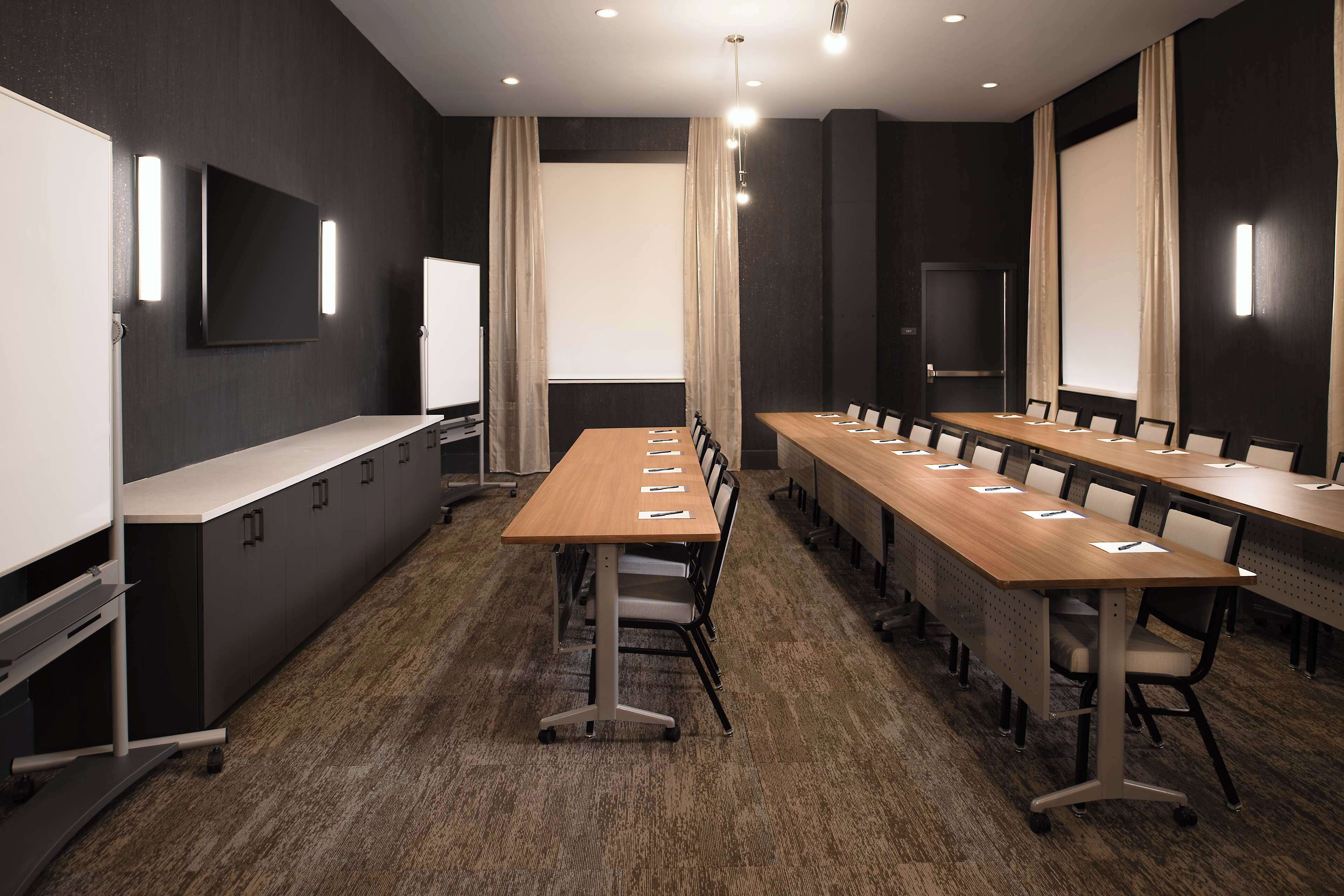 A conference room with long tables, chairs, a whiteboard, and a projector screen.
