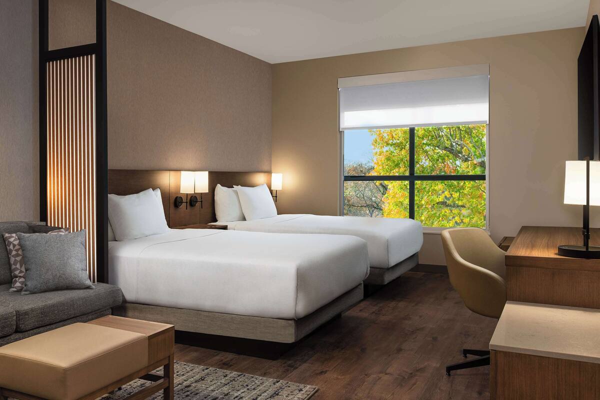 A hotel room with two beds, a sofa, a desk, and a large window with a view of autumn foliage.