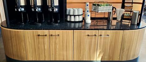 A coffee station with three coffee dispensers, a stack of white cups, and a Tazo coffee display.
