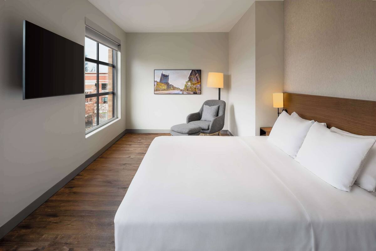 A hotel room with a large bed, a chair, a flat-screen TV mounted on the wall, and a painting of a cityscape.