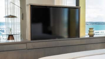 Quarto moderno com uma televisão de tela plana grande presa a uma parede de madeira, uma mesinha de cabeceira com luminária e vista para o mar por grandes janelas.