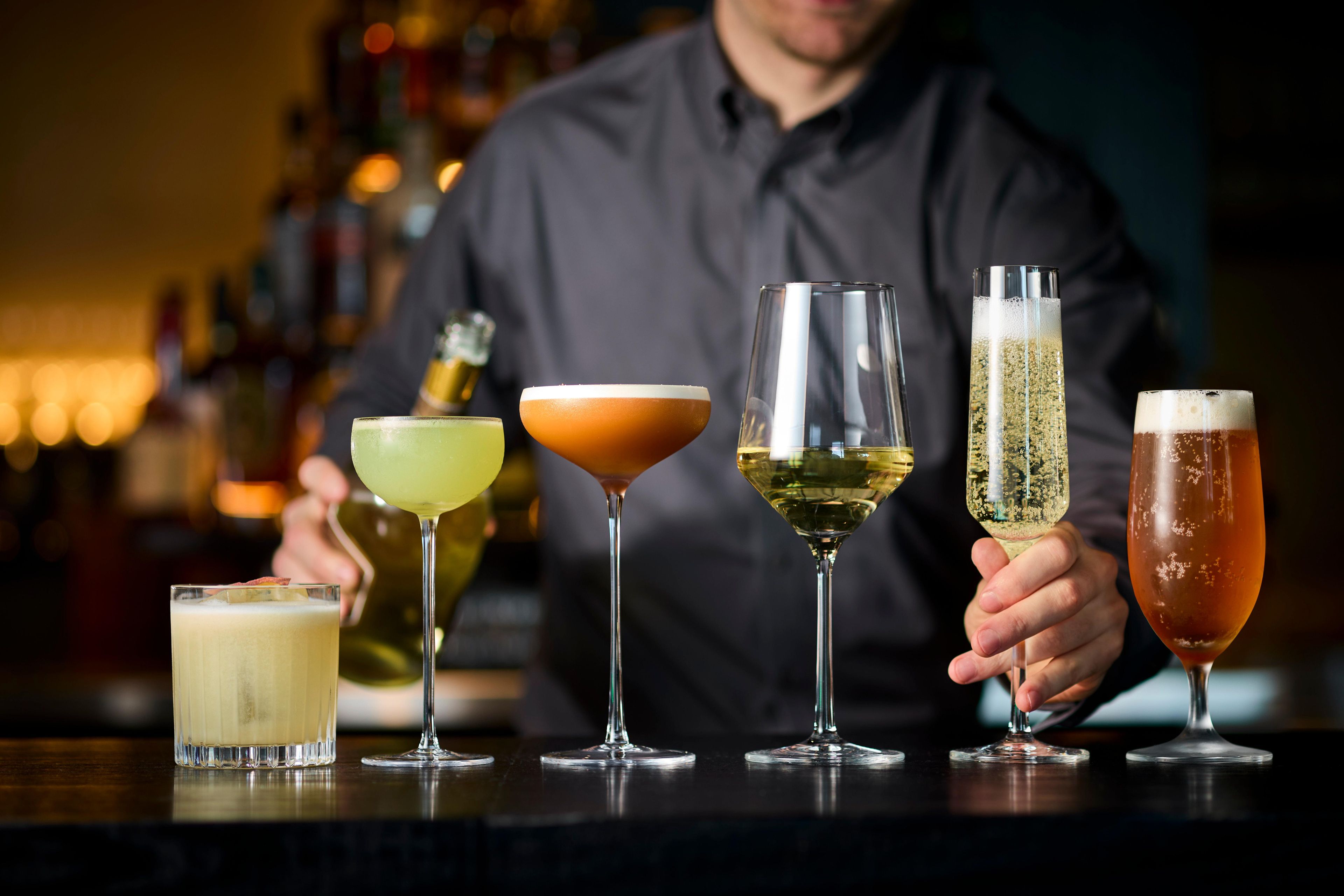 A bartender holding a glass of champagne, with various cocktails on the bar counter.