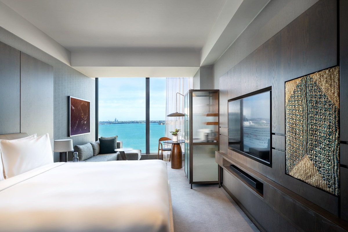 Quarto de hotel moderno com uma cama grande, televisão, área de estar e vista para a água.
