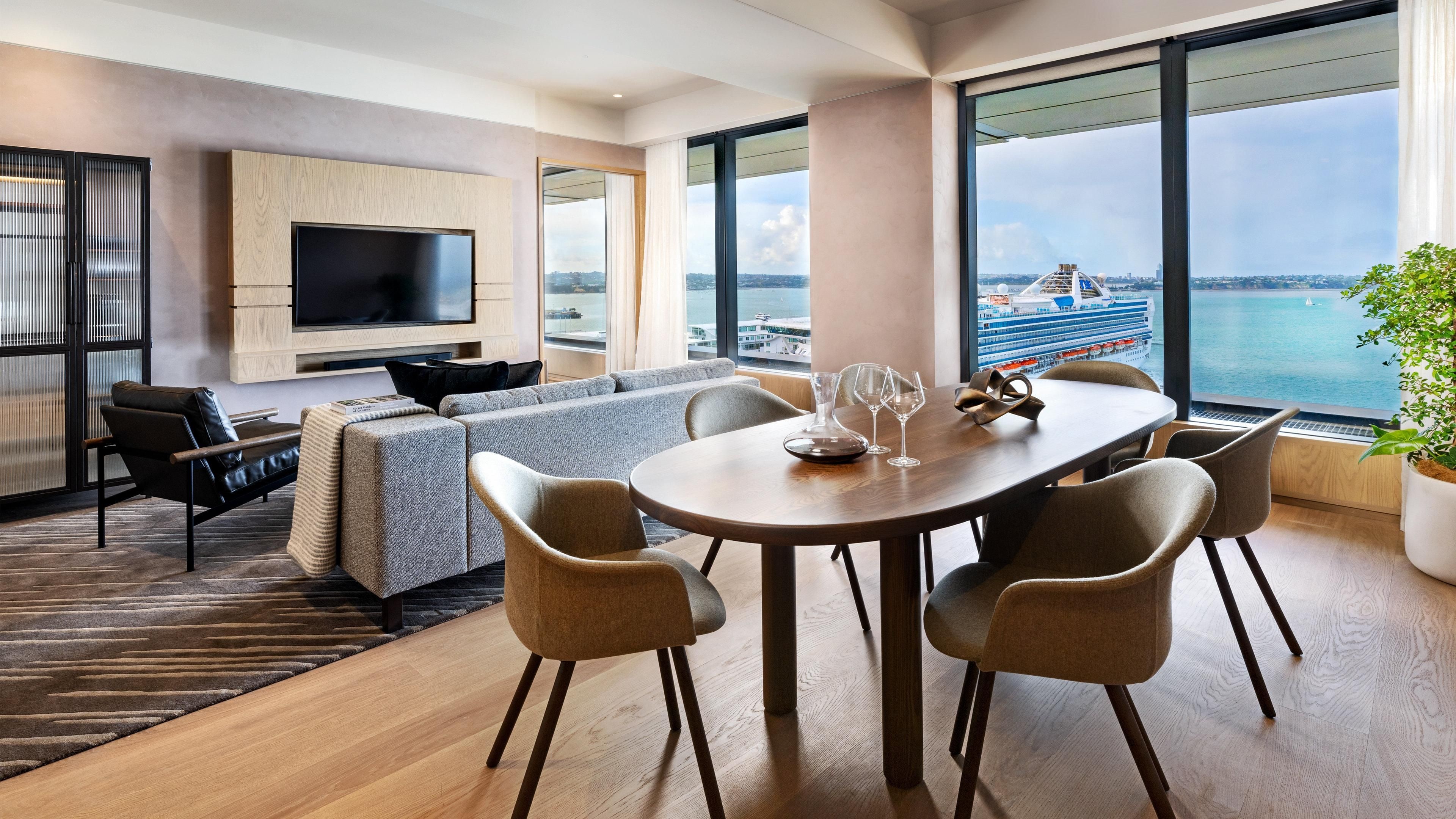 Una habitación de hotel moderna con un ventanal grande, un televisor de pantalla plana, un sofá, una mesa de comedor con sillas y una planta en maceta.