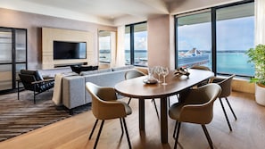 Quarto de hotel moderno com uma janela grande, televisão de tela plana, sofá, mesa de jantar com cadeiras e um vaso de planta.