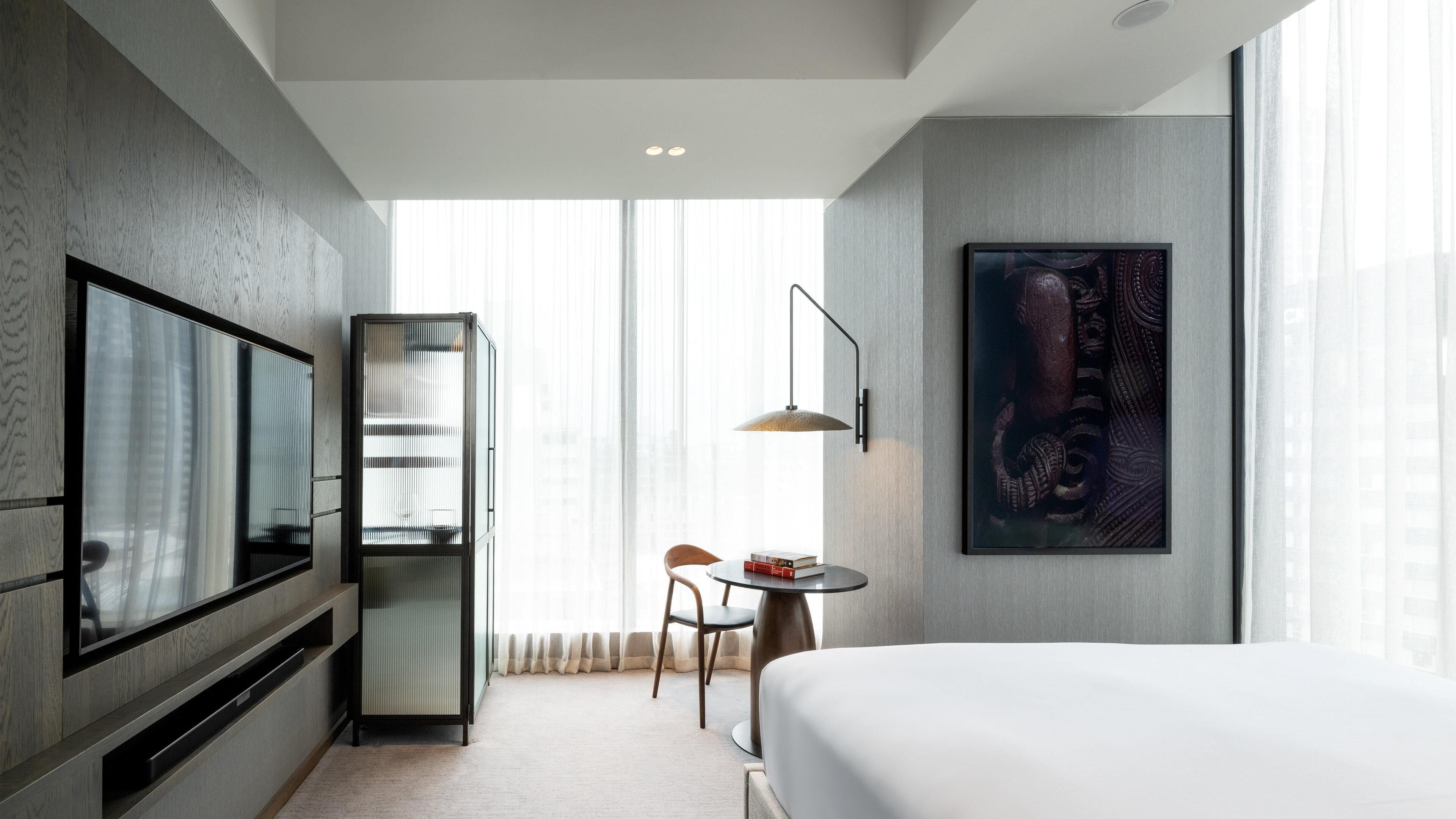 A hotel room with a bed, a TV, a desk with a chair, a bookshelf, and a large window.
