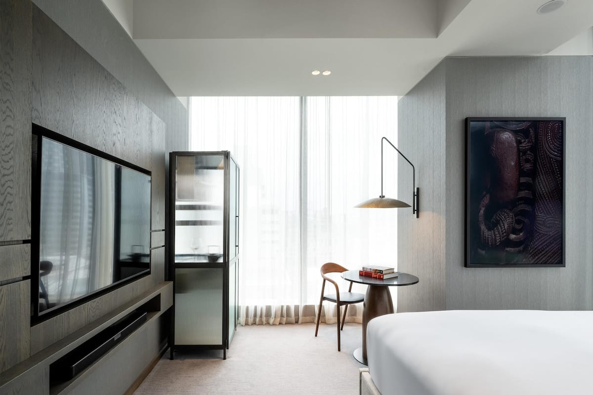 A hotel room with a bed, a TV, a desk with a chair, a bookshelf, and a large window.