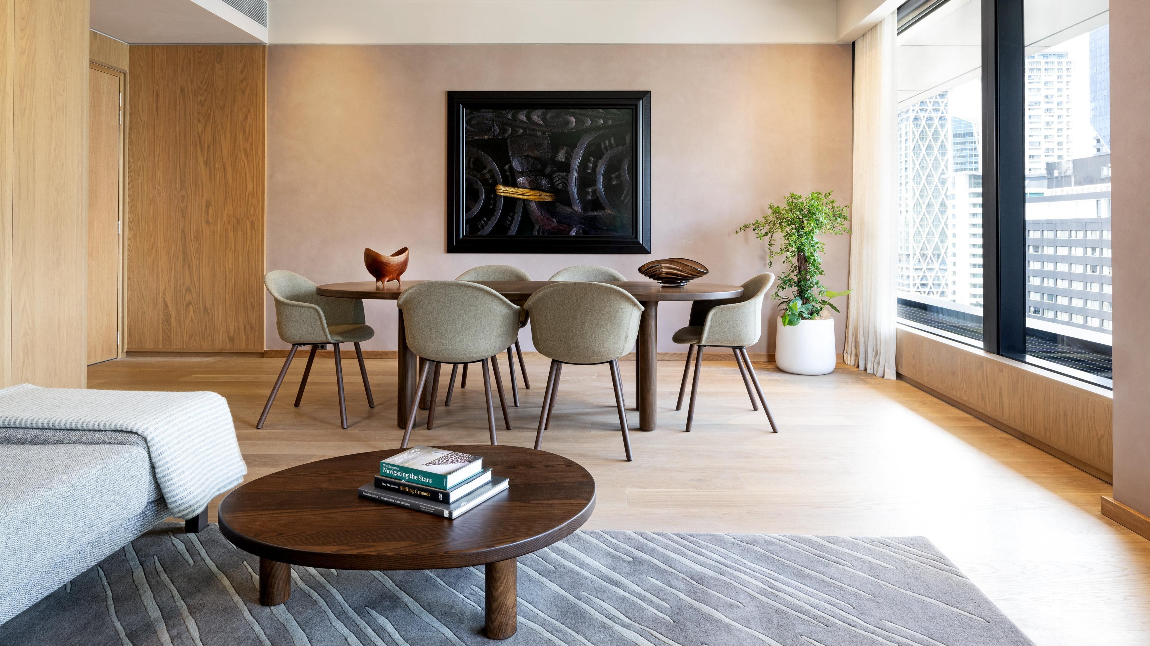 A modern living room with a round coffee table, a dining table with chairs, a large window with a city view, and a framed artwork on the wall.