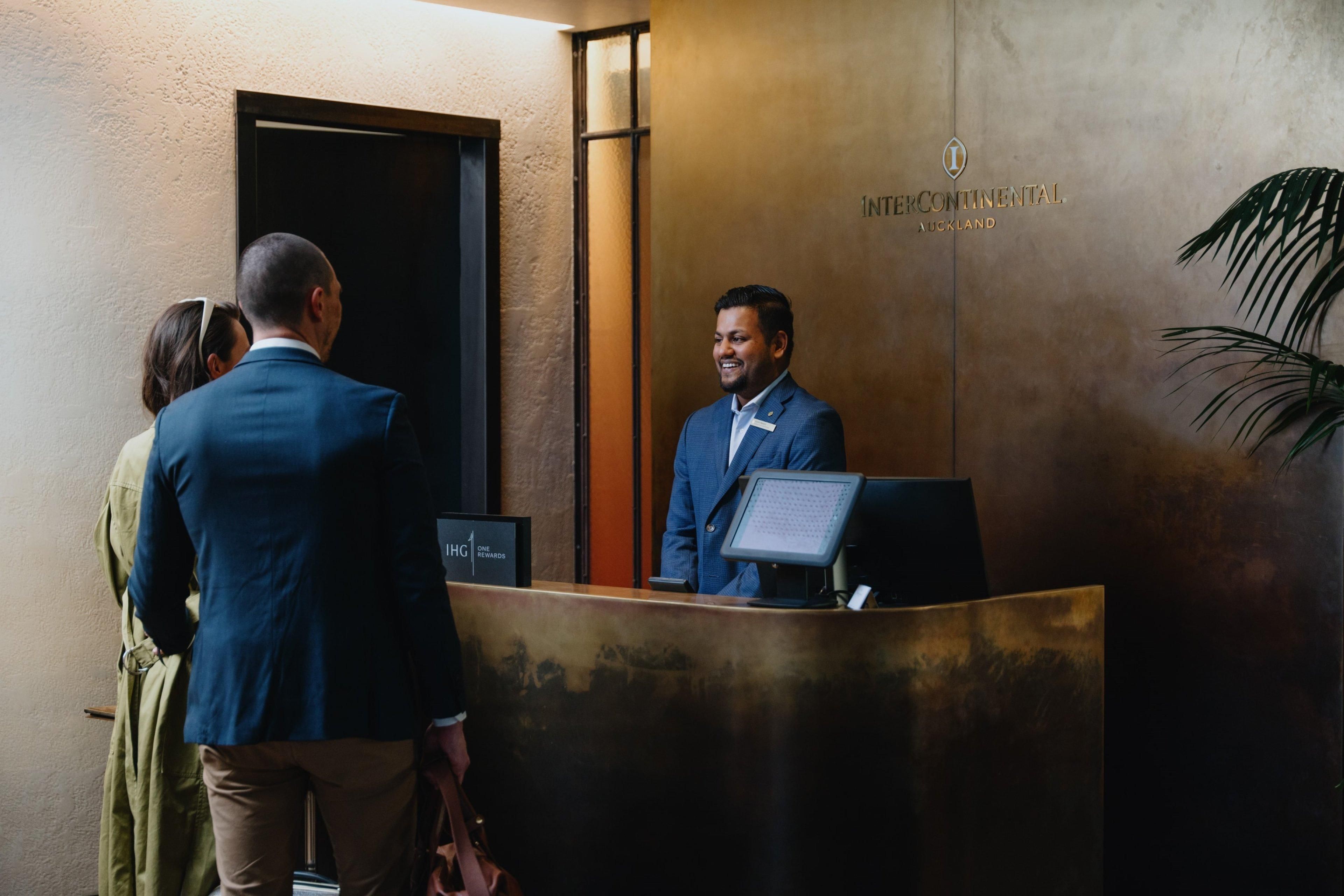 Eine Rezeption mit einem Mann im Anzug, einer Frau im grünen Kleid und einem weiteren Mann im blauen Anzug. An der Wand befindet sich ein Schild mit der Aufschrift 'INTERCONTINENTAL, AUCKLAND.'