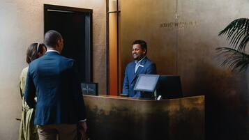 Eine Rezeption mit einem Mann im Anzug, einer Frau im grünen Kleid und einem weiteren Mann im blauen Anzug. An der Wand befindet sich ein Schild mit der Aufschrift 'INTERCONTINENTAL, AUCKLAND.'