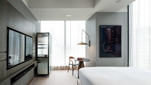 Habitació d'hotel amb un llit, una televisió, una escriptori amb una cadira, una prestatgeria i una finestra gran.