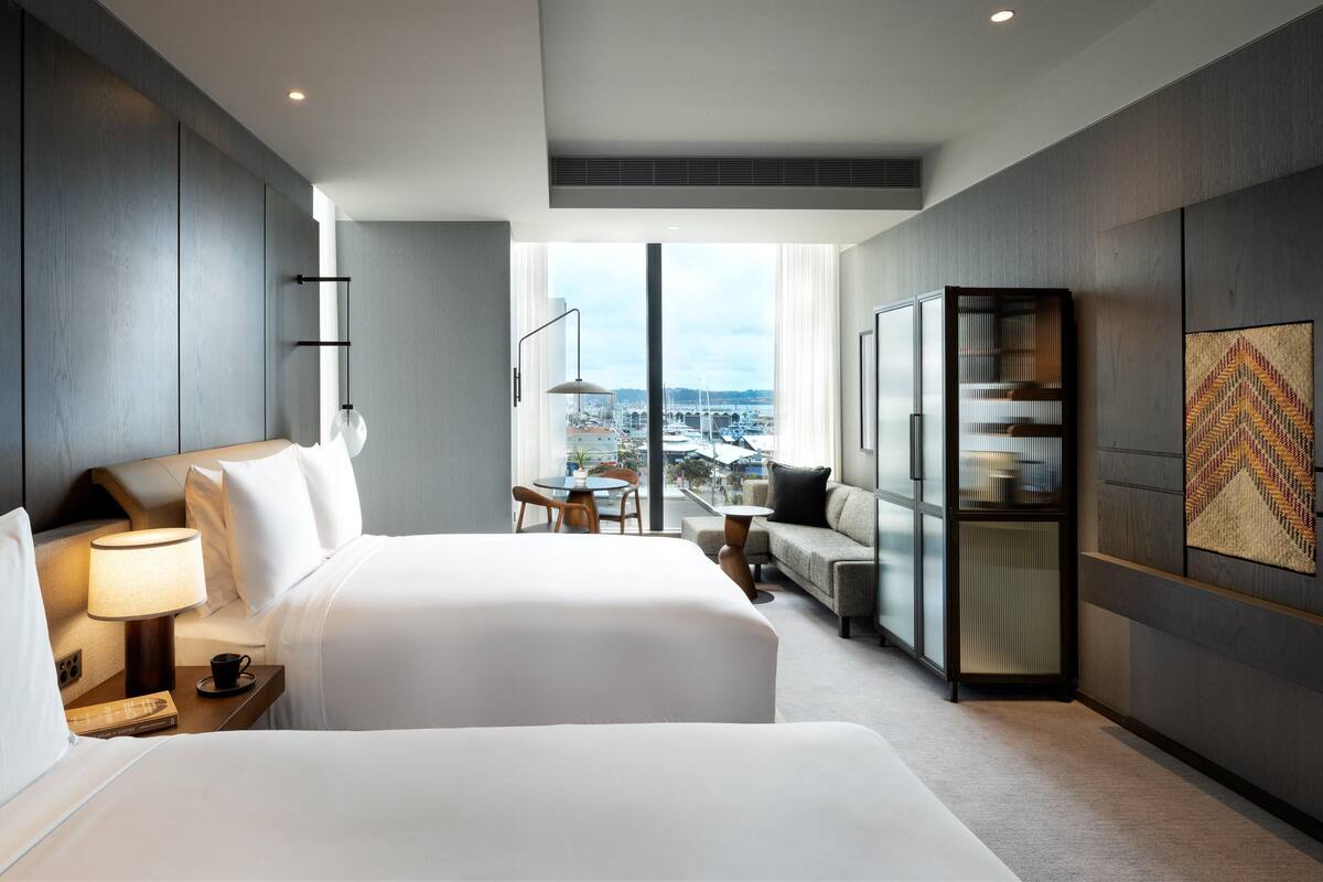 A modern hotel room with a large bed, bedside table, lamp, and a view of the city through a large window.