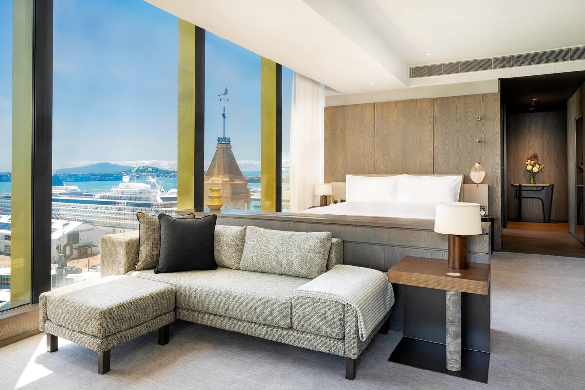 A modern hotel room with a large bed, a sofa, a small table, and a view of a cruise ship and cityscape.