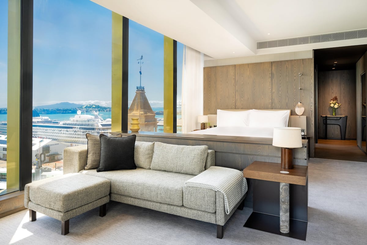 Une chambre d’hôtel moderne avec un grand lit, un canapé, une petite table et une vue sur un navire de croisière et sur le paysage urbain.