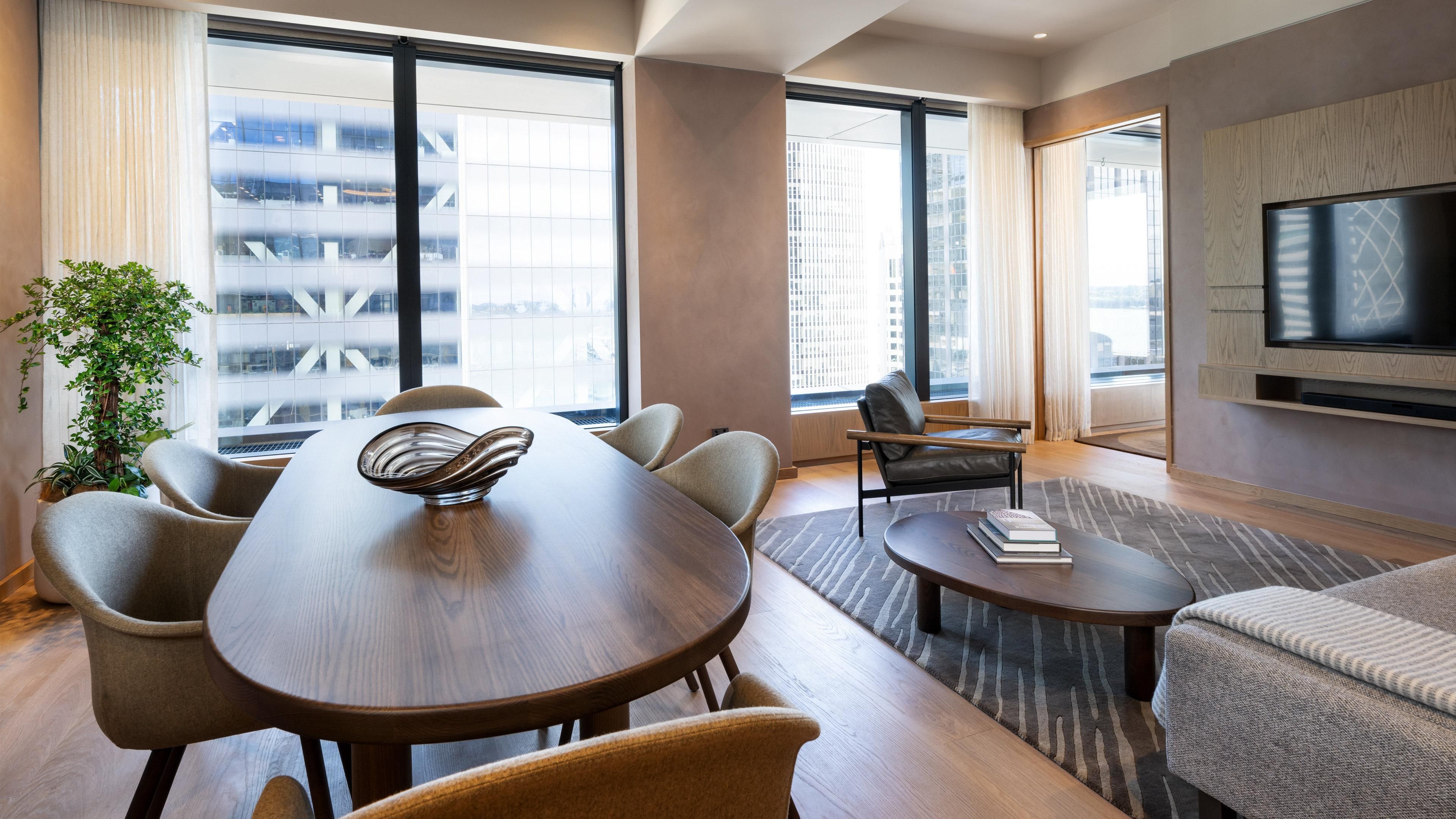 A modern hotel room with a dining table, chairs, a sofa, a TV, and large windows with a city view.