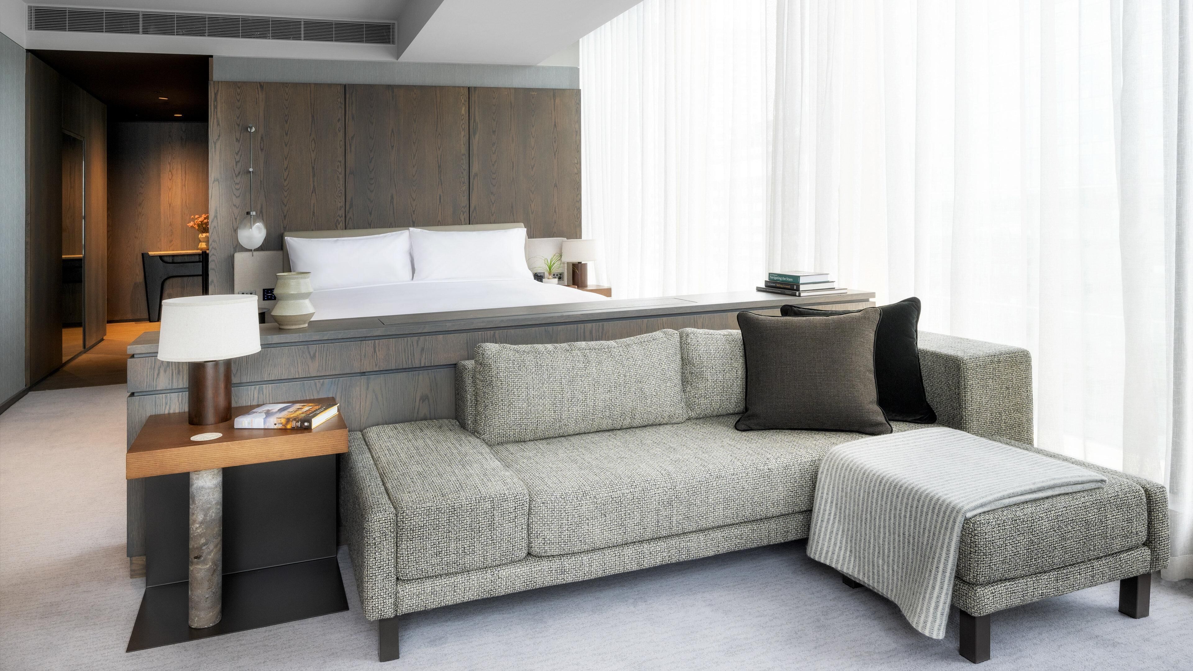 A modern hotel room with a bed, a sofa, a bedside table, and a lamp.