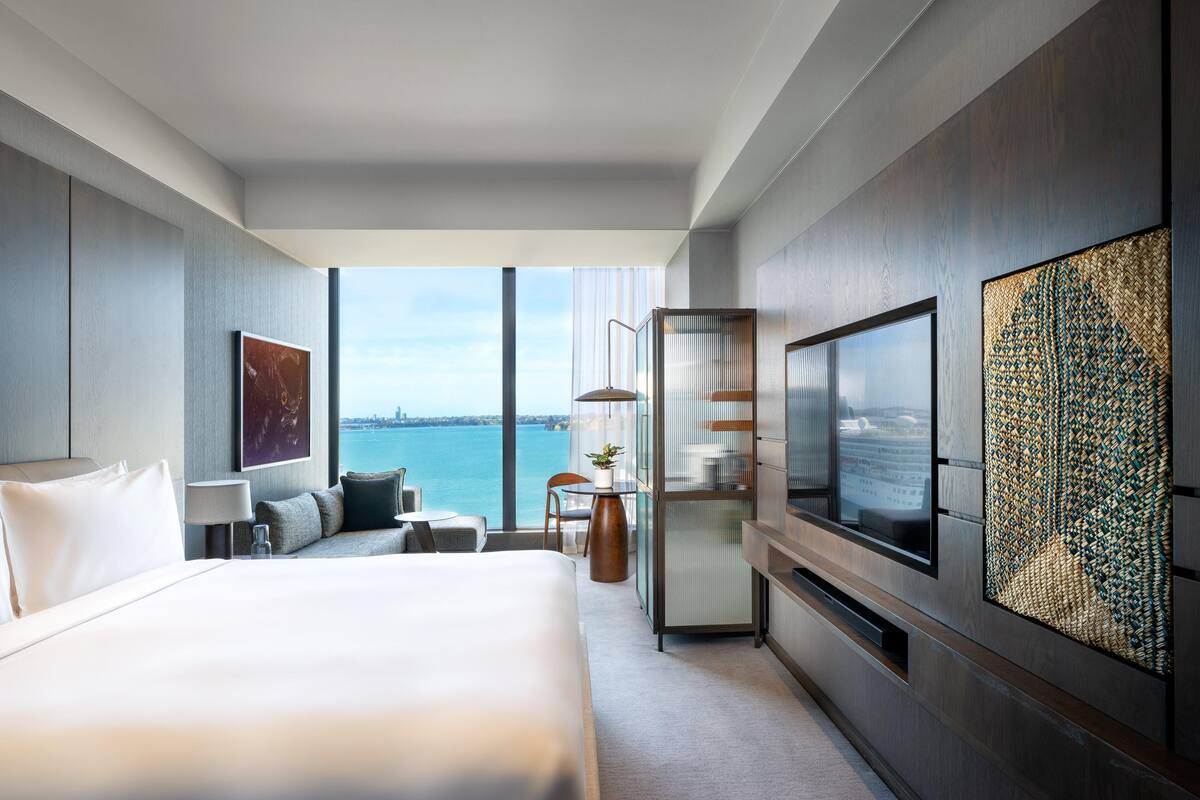 A modern hotel room with a large bed, a TV, a seating area, and a view of the water.
