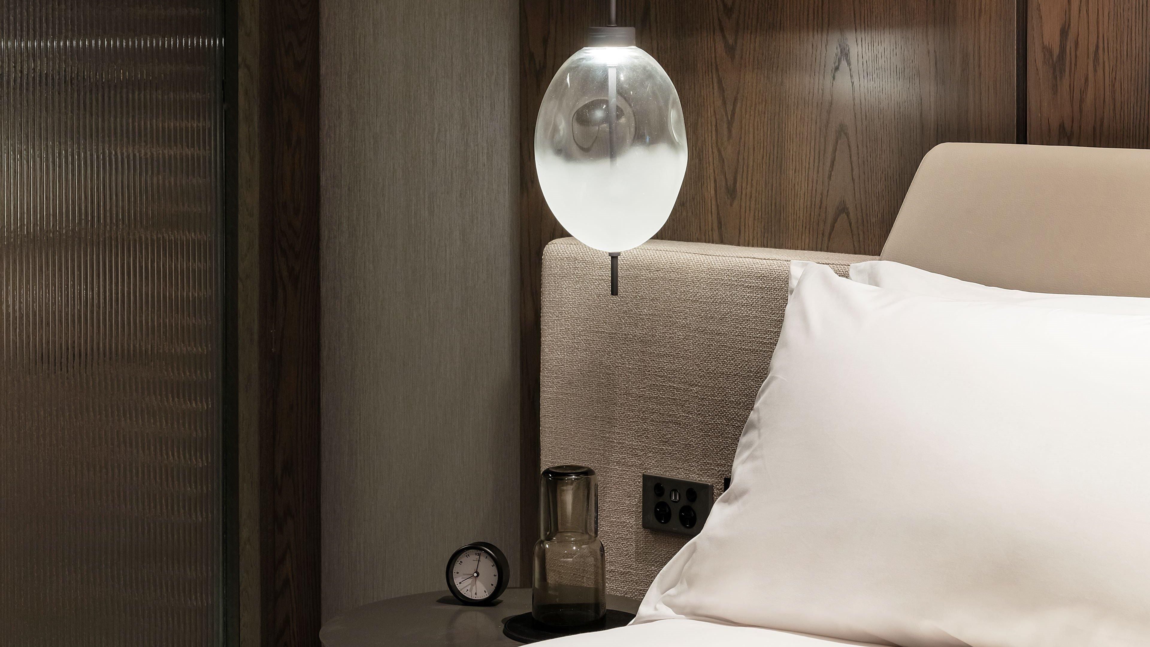 A hotel room with a bed, a bedside table with a lamp, and a wall clock.