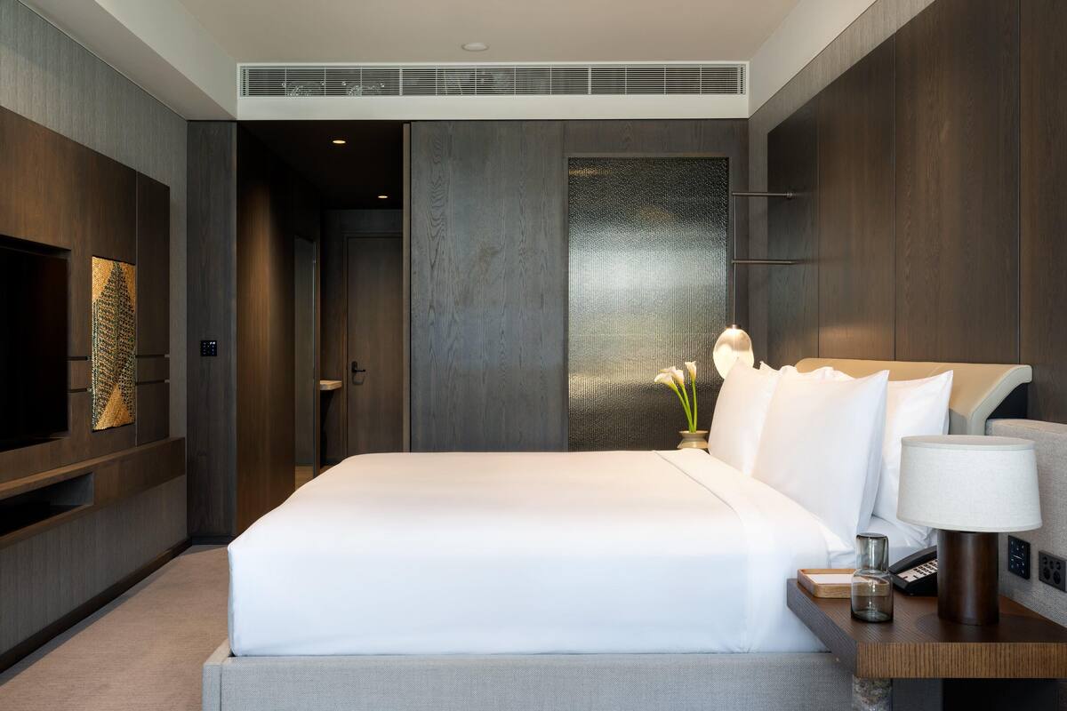 A modern hotel room with a large bed, a TV, a bedside table with a lamp, and a decorative wall panel.