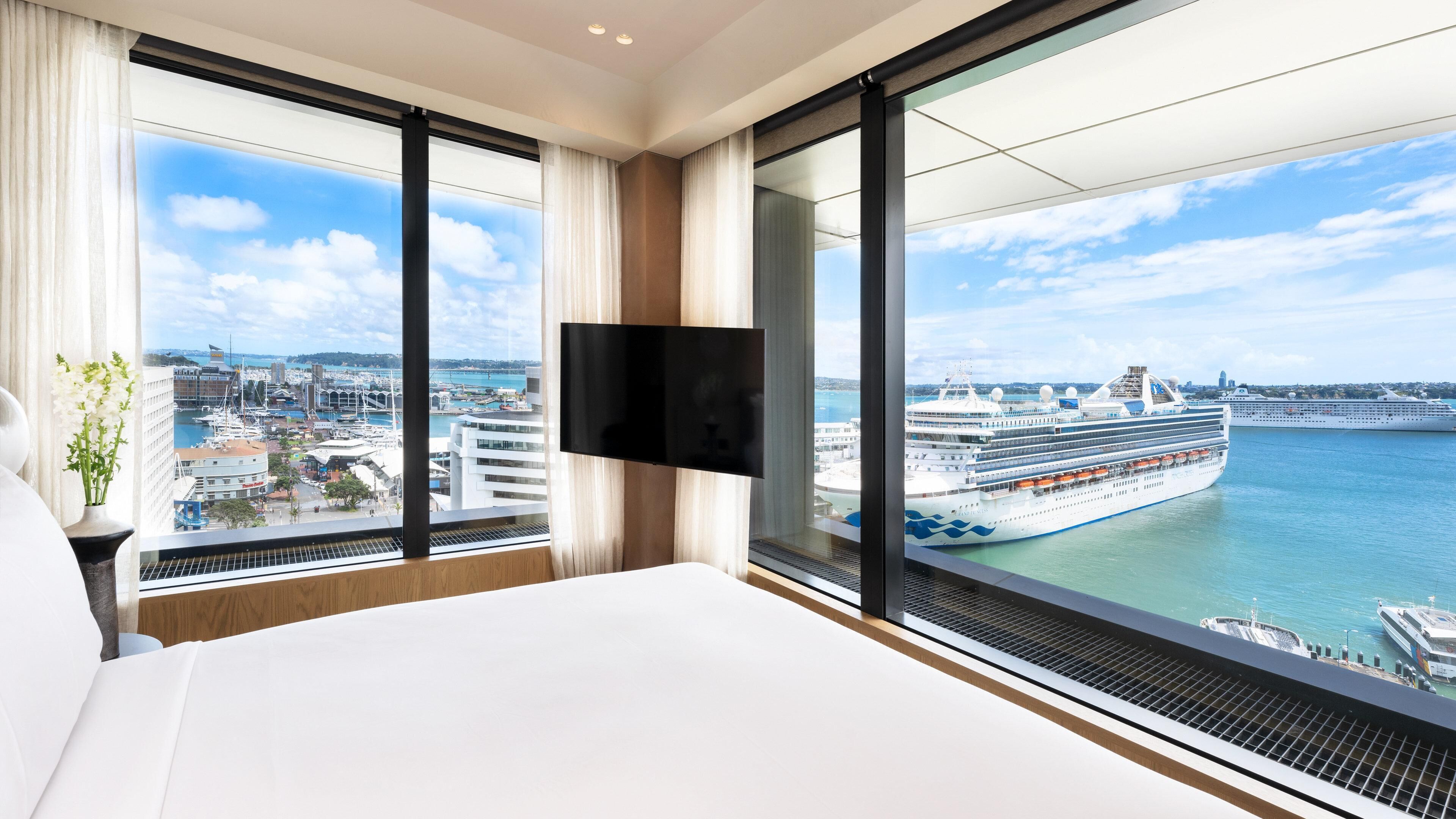 A hotel room with a large bed, a flat-screen TV mounted on the wall, and a view of a cruise ship docked at a port.