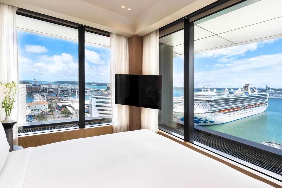 A hotel room with a large bed, a flat-screen TV mounted on the wall, and a view of a cruise ship docked at a port.