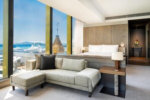 Habitació d'hotel moderna amb un llit gran, un sofà, una taula petita i vista d'un vaixell de creuer i un paisatge urbà.