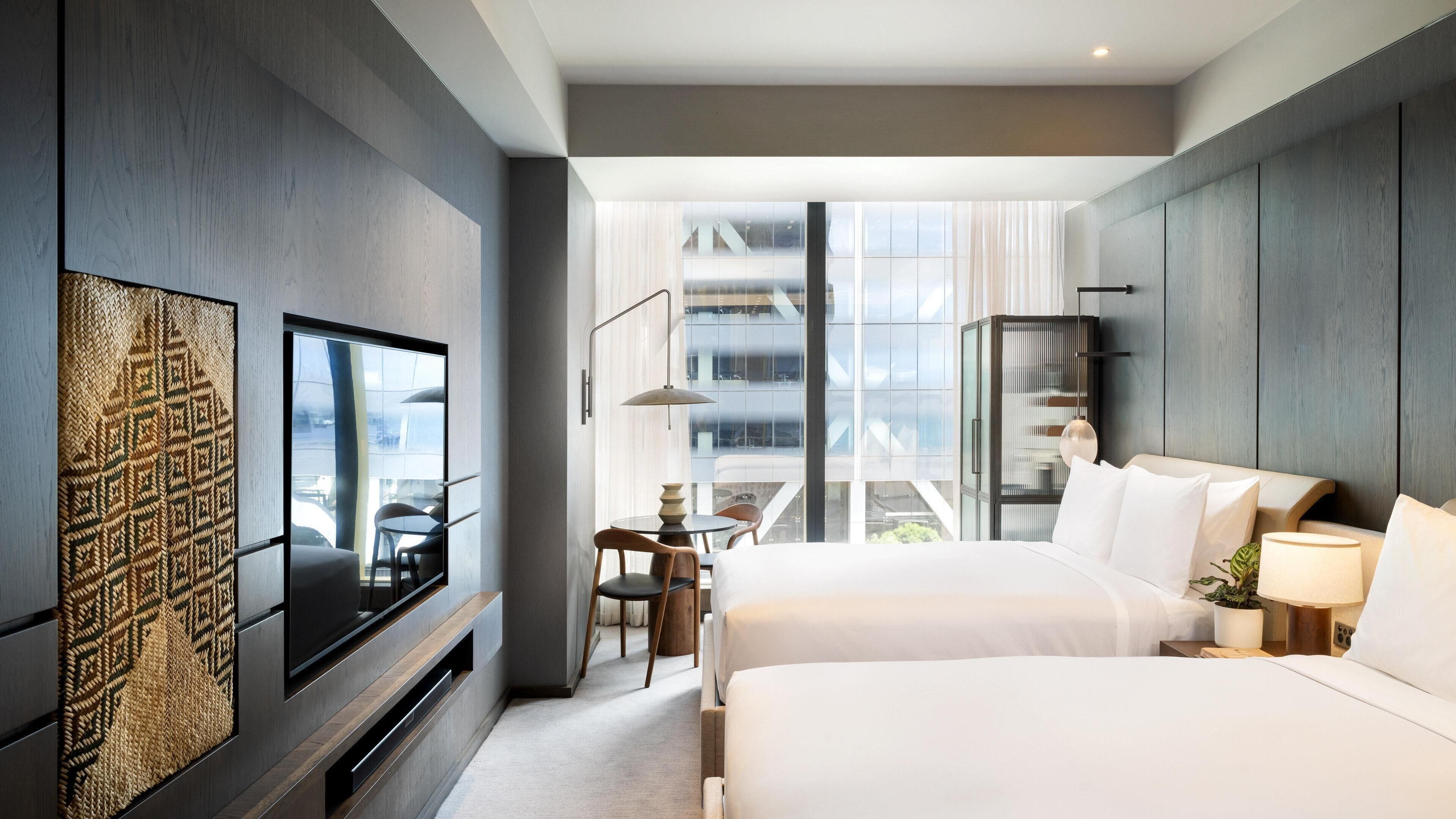 A modern hotel room with a large bed, a TV mounted on the wall, a dining area with a table and chairs, and a view of a building through large windows.