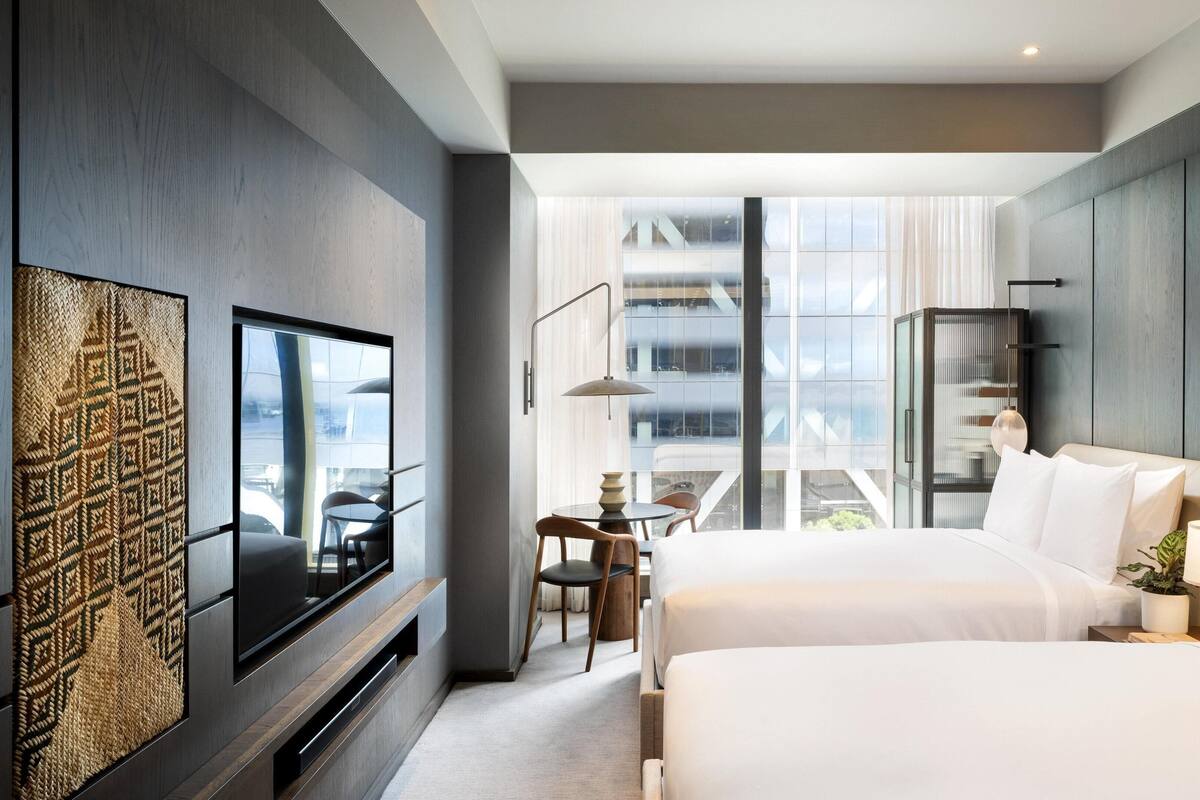 A modern hotel room with a large bed, a TV mounted on the wall, a dining area with a table and chairs, and a view of a building through large windows.