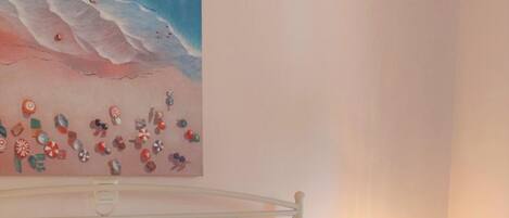 A neatly made bed with white linens and a rolled towel, a bedside table with a lamp, and a painting of a beach scene on the wall.