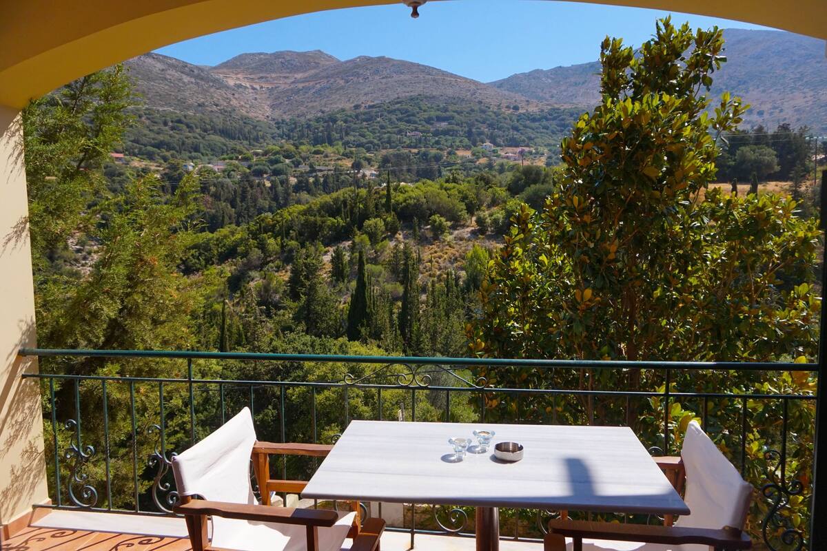 Ein Balkon mit Tisch und Stühlen, mit Blick auf eine gebirgige Landschaft mit Bäumen und Gebäuden.