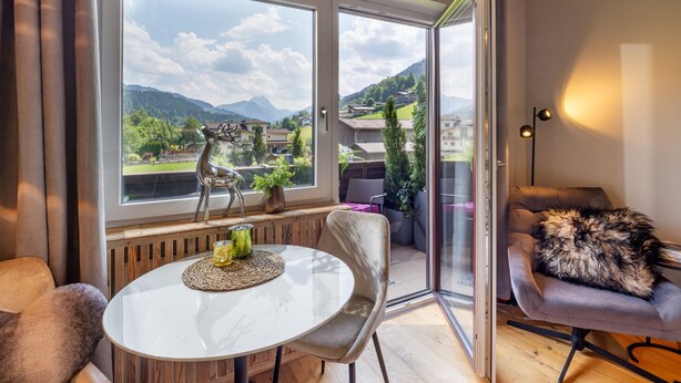 A modern living room with a glass door leading to a balcony, a round table with a placemat, a plush armchair, and a decorative deer sculpture. - Alpine's Nest studio apartment with mountain views (Kirchberg in Tirol)