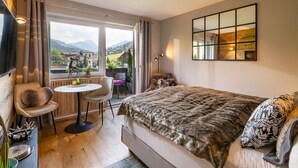 A bedroom with a large bed, a small round table, a chair, a sofa, and a TV. There is a window with a view of mountains and houses. - Alpine's Nest studio apartment with mountain views (Kirchberg in Tirol)