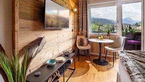 A modern room with a wooden wall, a flat-screen TV, a dining table with chairs, and a large window offering a mountain view. - Alpine's Nest studio apartment with mountain views (Kirchberg in Tirol)
