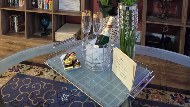A cozy living room with a fireplace, bookshelves, a TV, and a table set with flowers, champagne, and a menu.