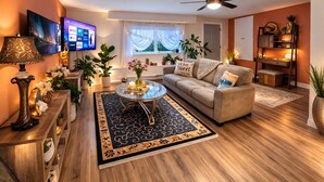 A living room with a sofa, coffee table, and TV. There are plants, a lamp, and a bookshelf.