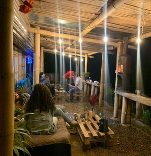 Uma área de estar externa com pilares de bambu, lareira a lenha e luminárias penduradas.