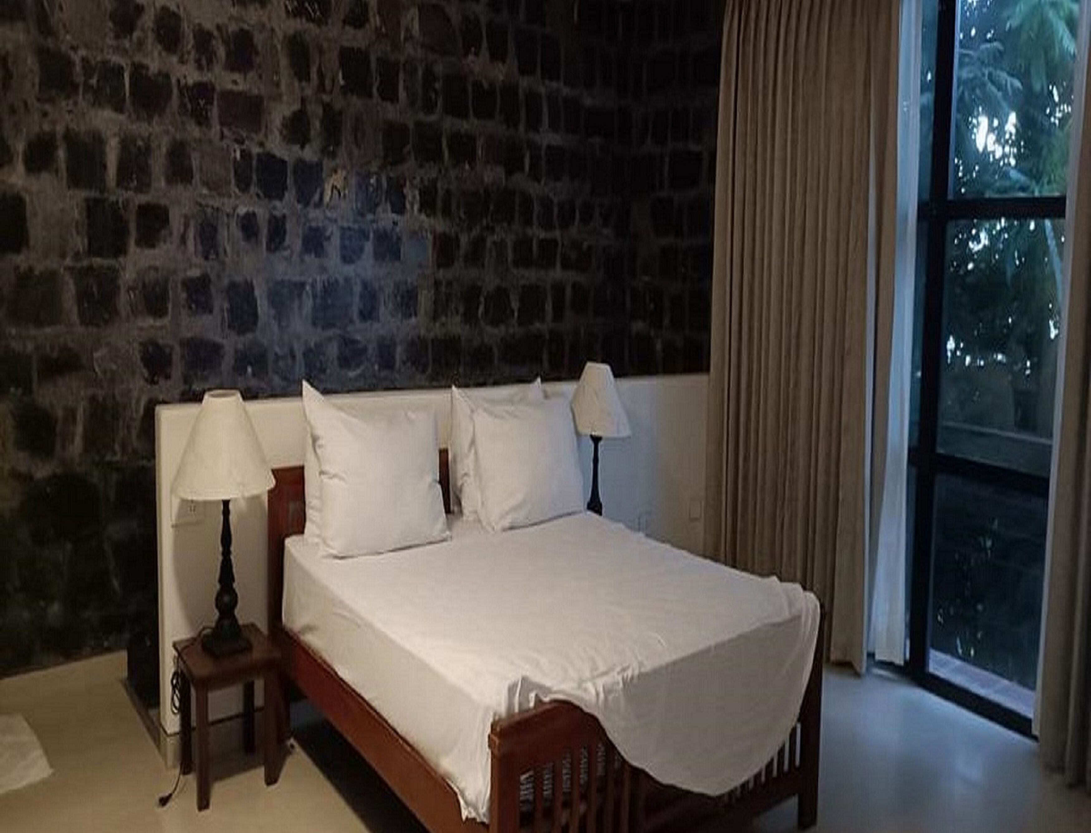 Un dormitorio con pared de piedra, una cama con ropa de cama blanca, dos mesitas de noche con lámparas y un ventanal con cortinas.