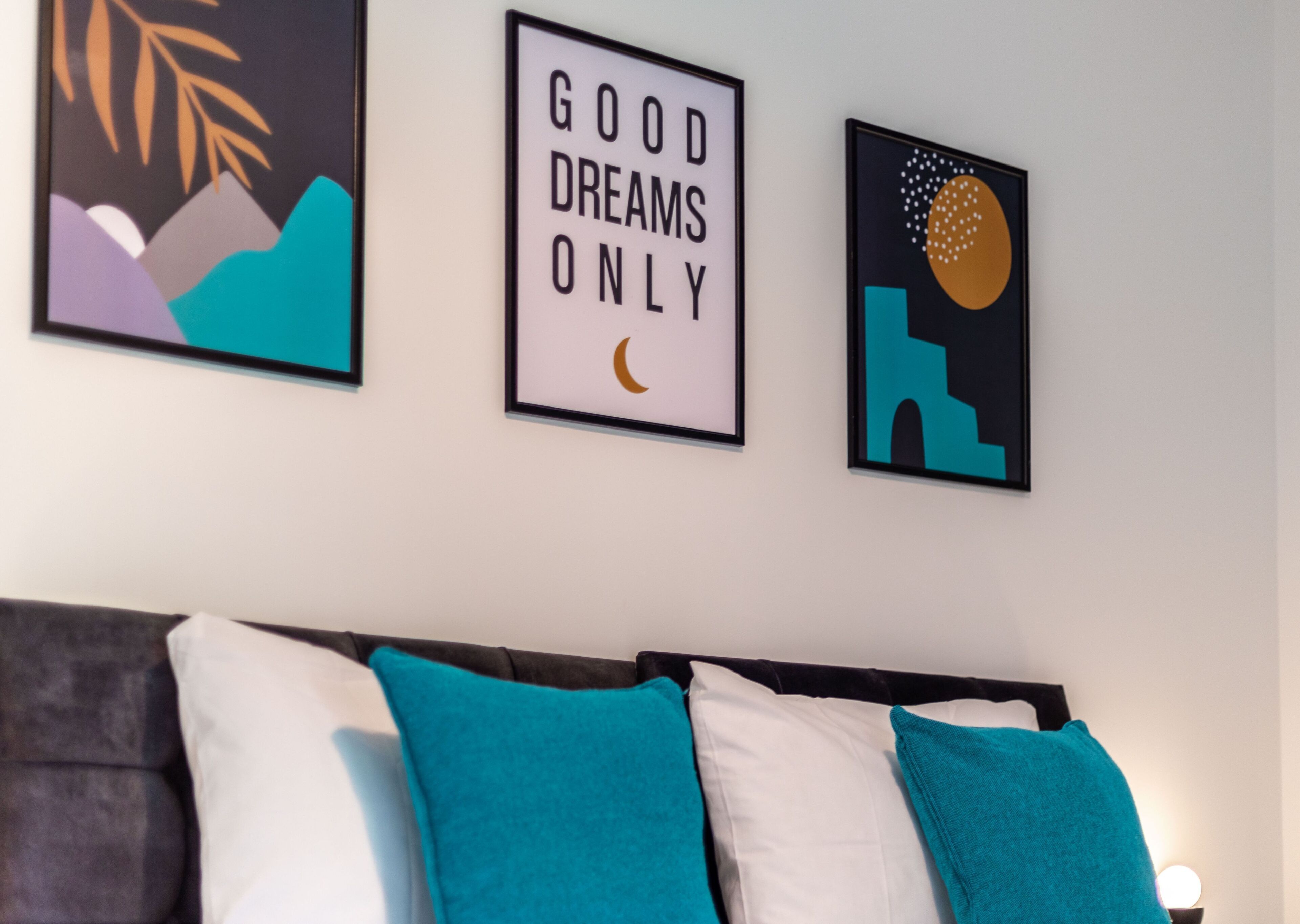 A bedroom with a bed, teal and white pillows, and three framed artworks on the wall.
