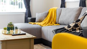 Una sala de estar moderna con un sofá gris, una manta amarilla y una pequeña mesa con plantas en macetas y un control remoto.