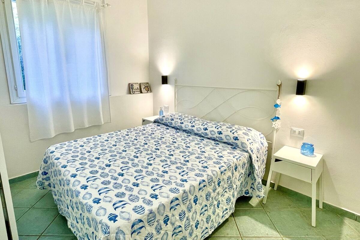 Un dormitorio con cama, mesita de noche, lámpara de pared y ventana con cortinas.