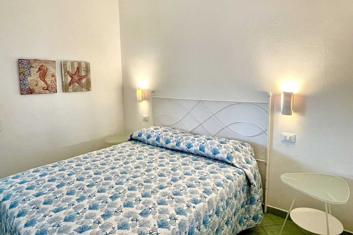 Un dormitorio con una cama, dos luces fijadas a la pared, una mesita blanca y dos obras de arte colgadas.