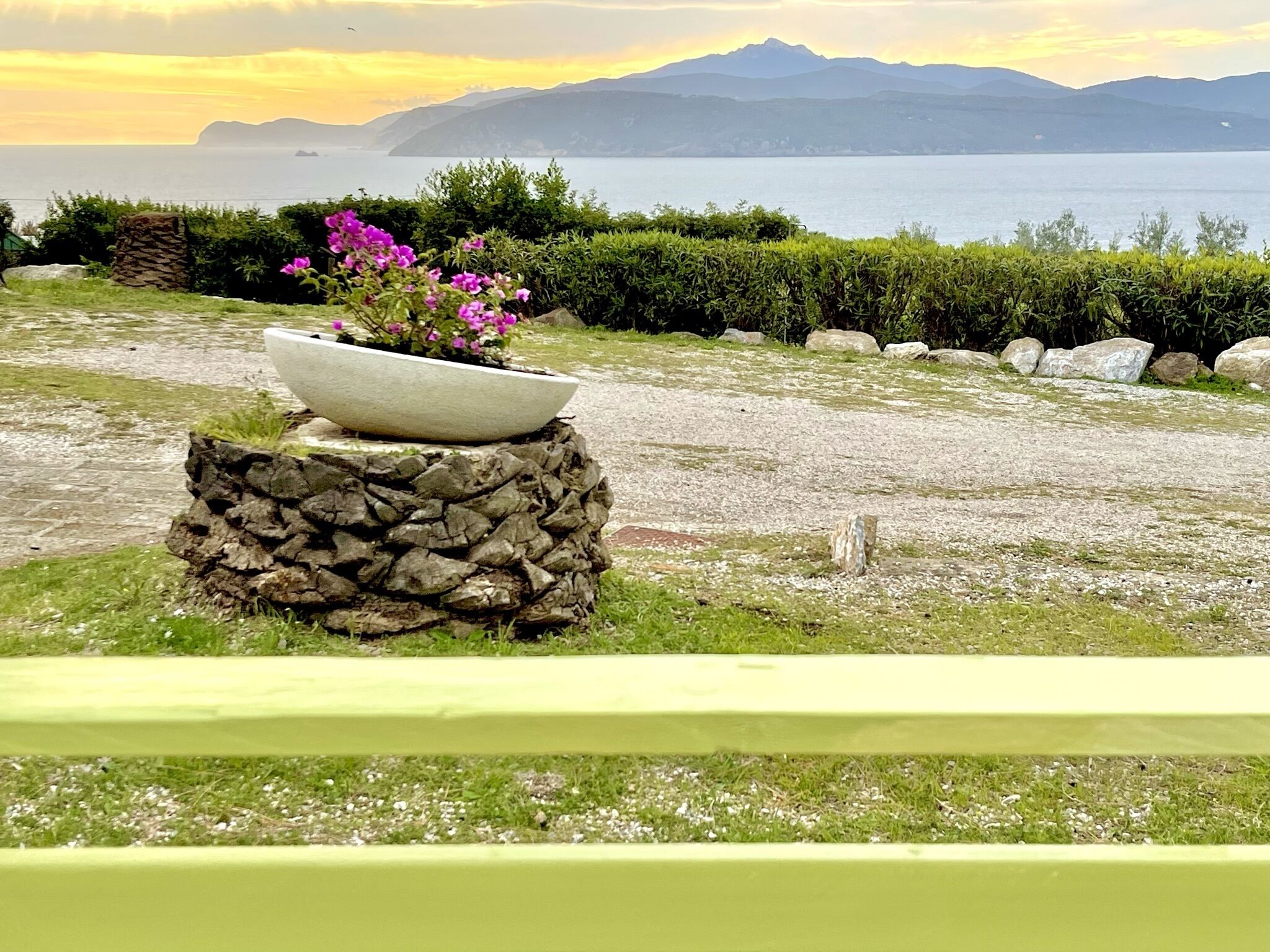 A garden with a stone planter holding pink flowers, a gravel path, and a view of the sea and mountains in the background.