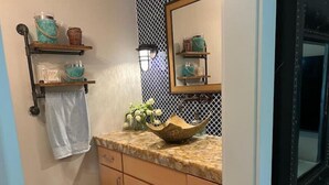 Une salle de bain avec un comptoir de couleur beige, un lavabo orné d’un récipient décoratif, un miroir et des étagères contenant des articles de toilette.
