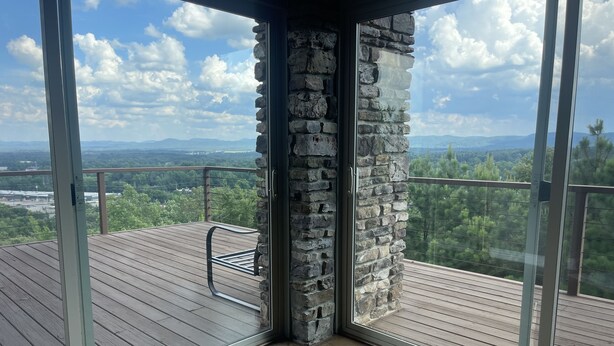 Een balkon met een stenen pilaar, een houten terras en glazen deuren met uitzicht op een bergachtig landschap.
