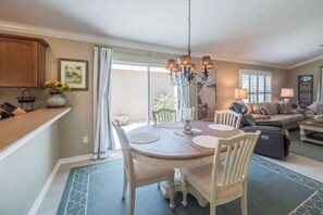 A dining area with a round table, chairs, and a chandelier. There is a sofa and a window with blinds in the background.
