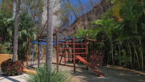 A playground with climbing structures and a slide, surrounded by palm trees and plants. - Beautiful Condo with Private Beach Club (Cruz de Huanacaxtle)