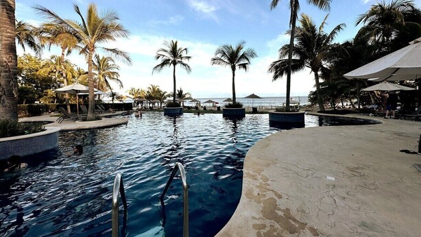 A swimming pool with palm trees and umbrellas, surrounded by a concrete deck. - Beautiful Condo with Private Beach Club (Cruz de Huanacaxtle)