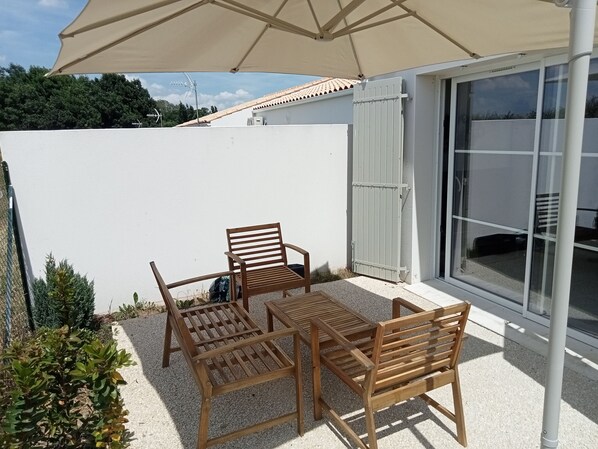 Een terras met een tafel en vier stoelen, een beige parasol en een schuifdeur met glas.